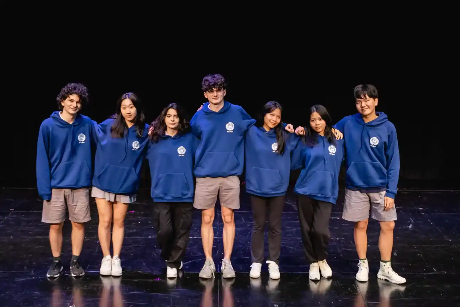 Enthusiastic diverse students wearing blue hoodies standing on stage at World Schools event.