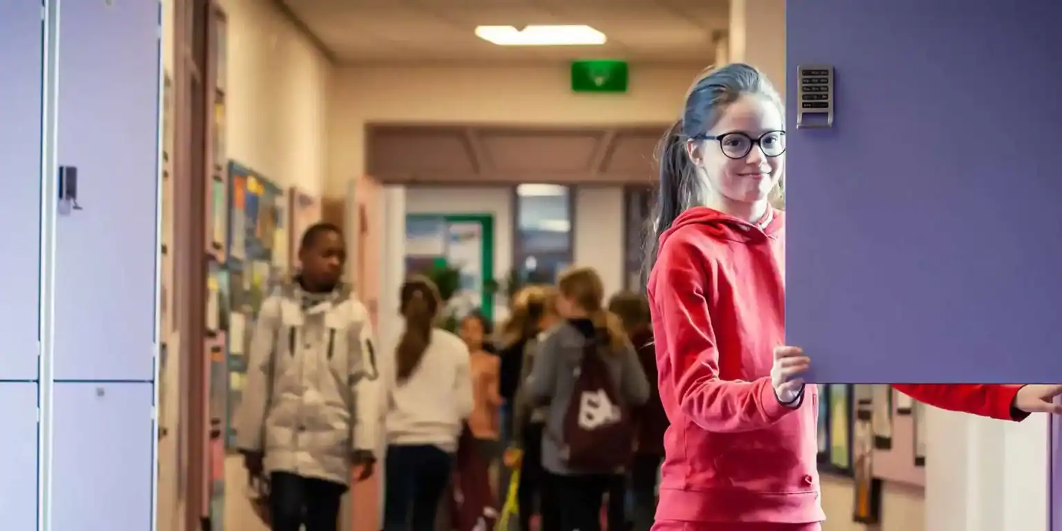 Jasna nastolatka w okularach stojąca w szkolnej sali, uśmiechająca się do kamery.
