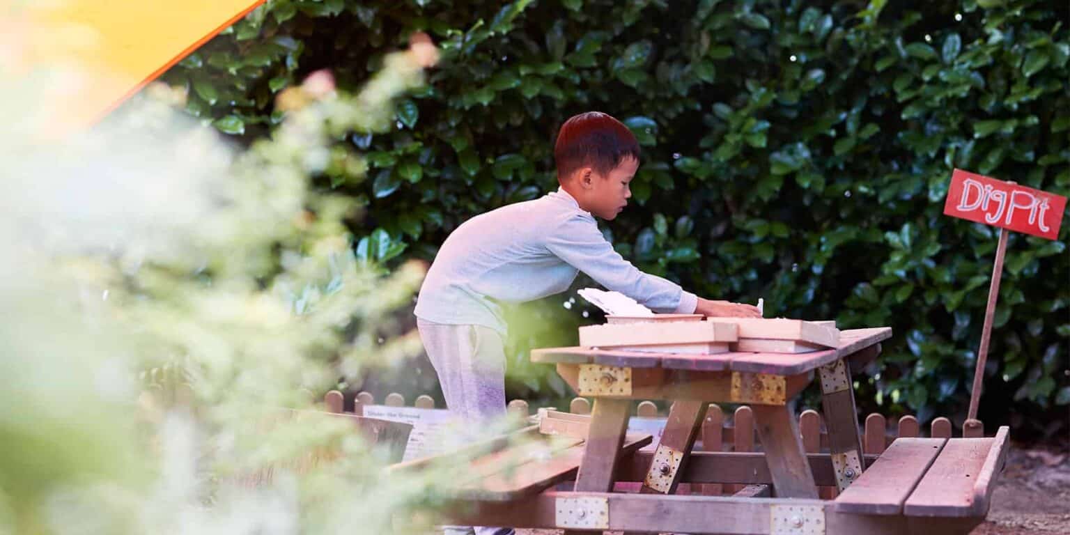 Child playing with dirt at an outdoor classroom, learning through hands-on activities in a nature-based school environment.