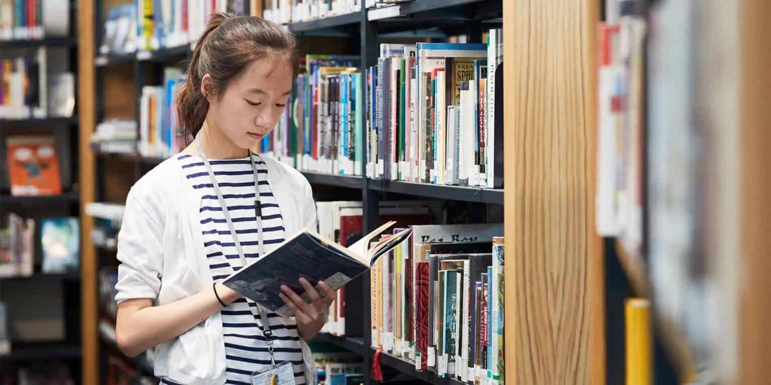 Young girl studying a book in a library with bookshelves in the background.