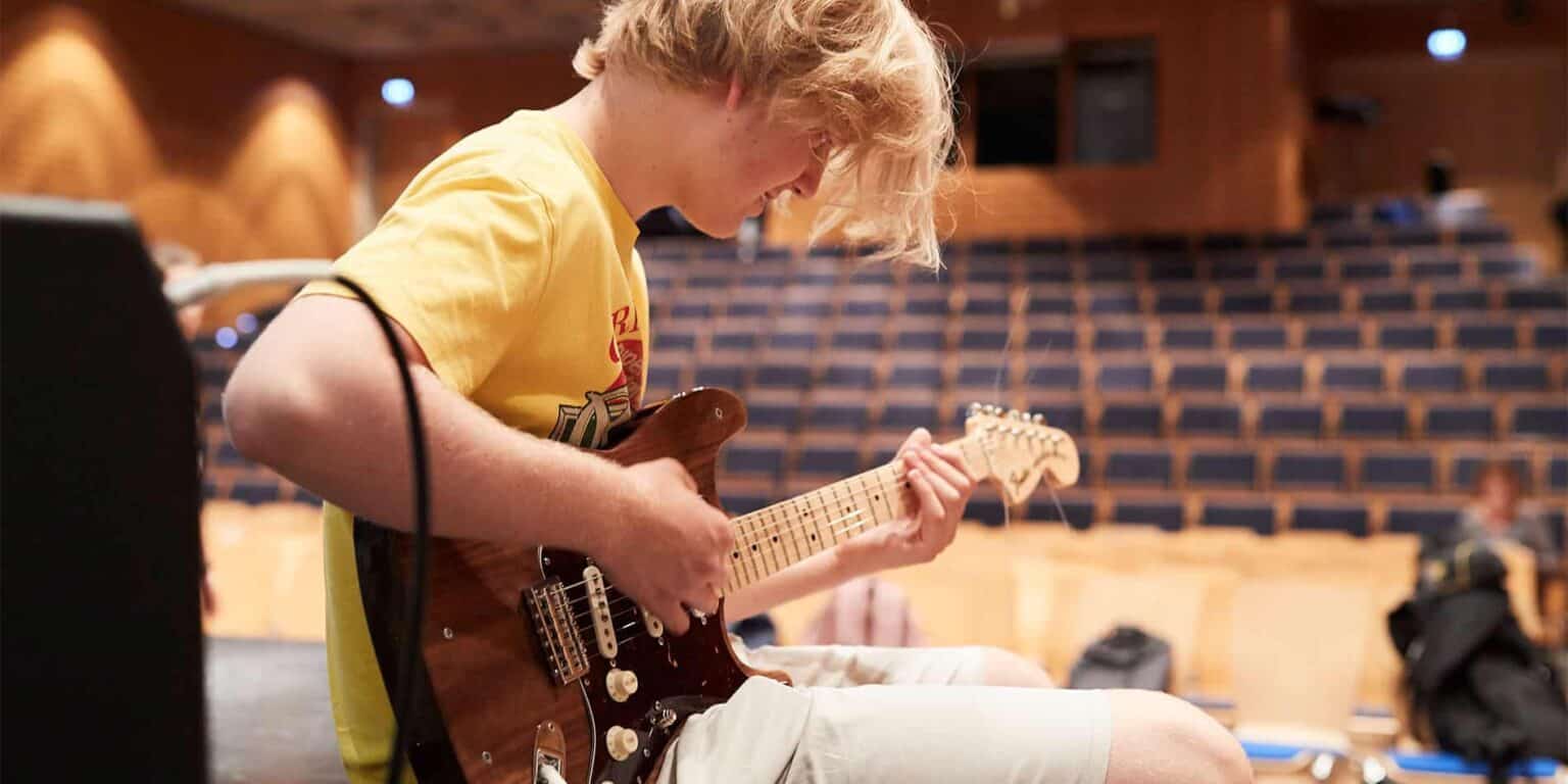 Innovative student playing electric guitar during school music class with tiered auditorium seating.