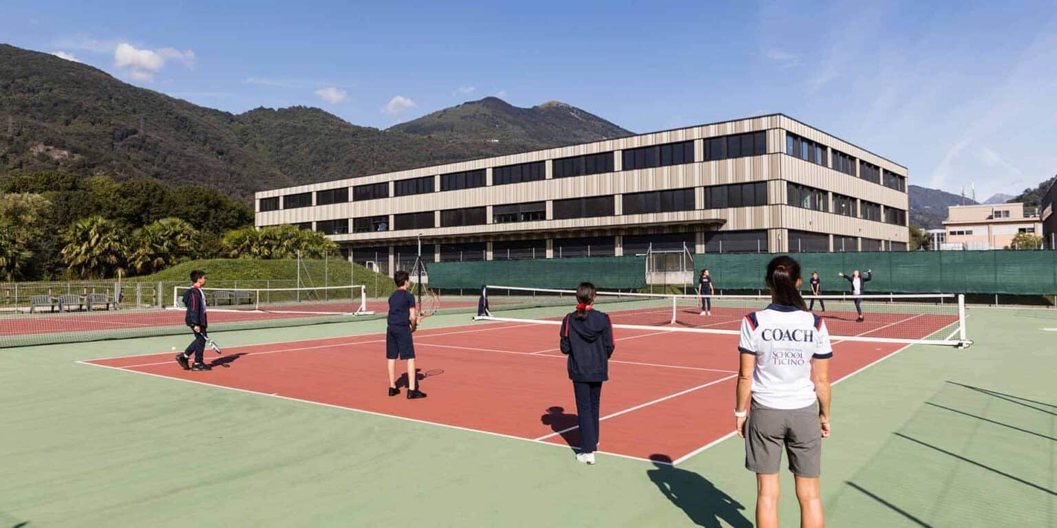Tenniscoaching op een moderne internationale school met bergen als achtergrond, met de nadruk op sport en buitenschoolse activiteiten.