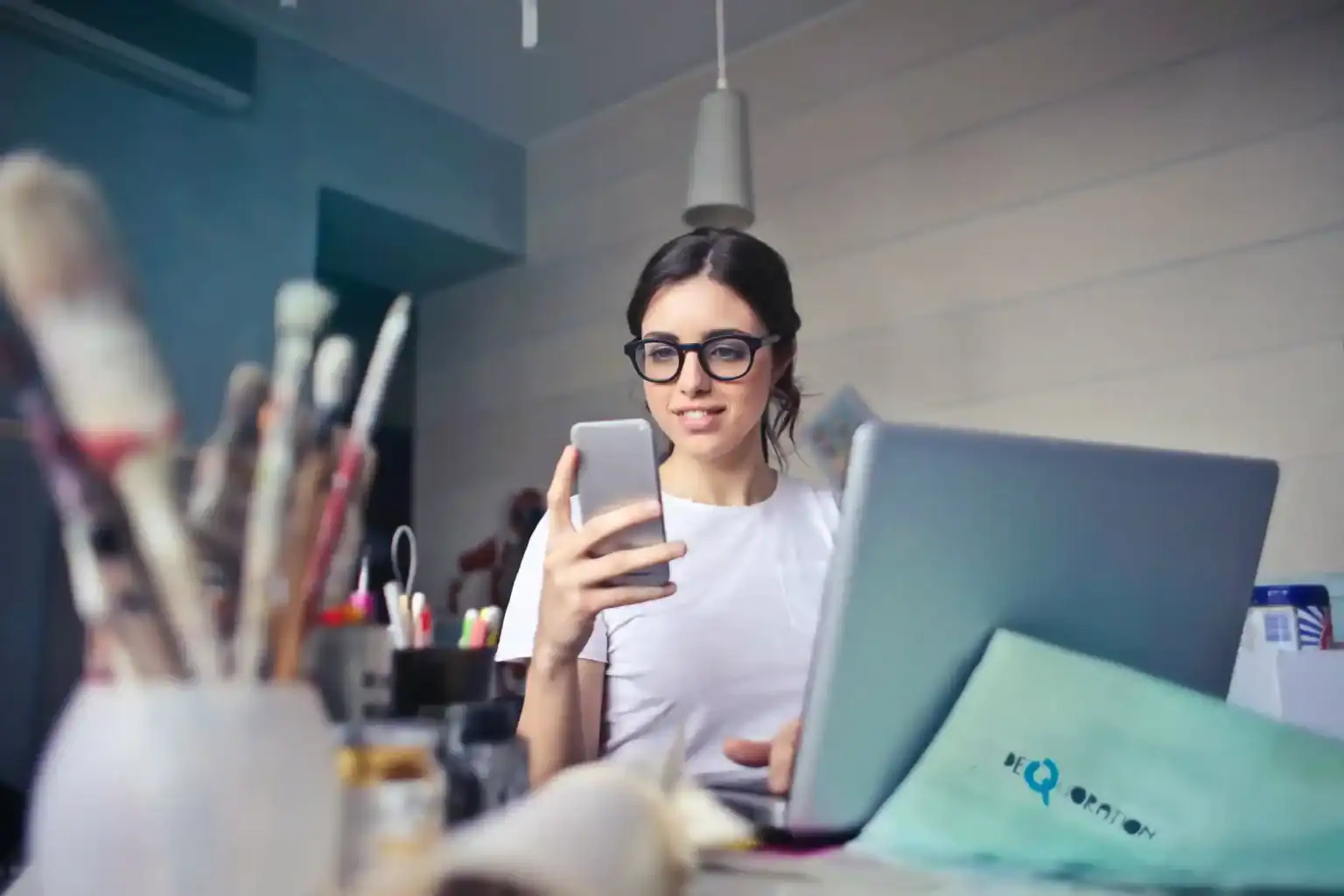 Bright female student using smartphone and laptop in modern study space with art supplies.