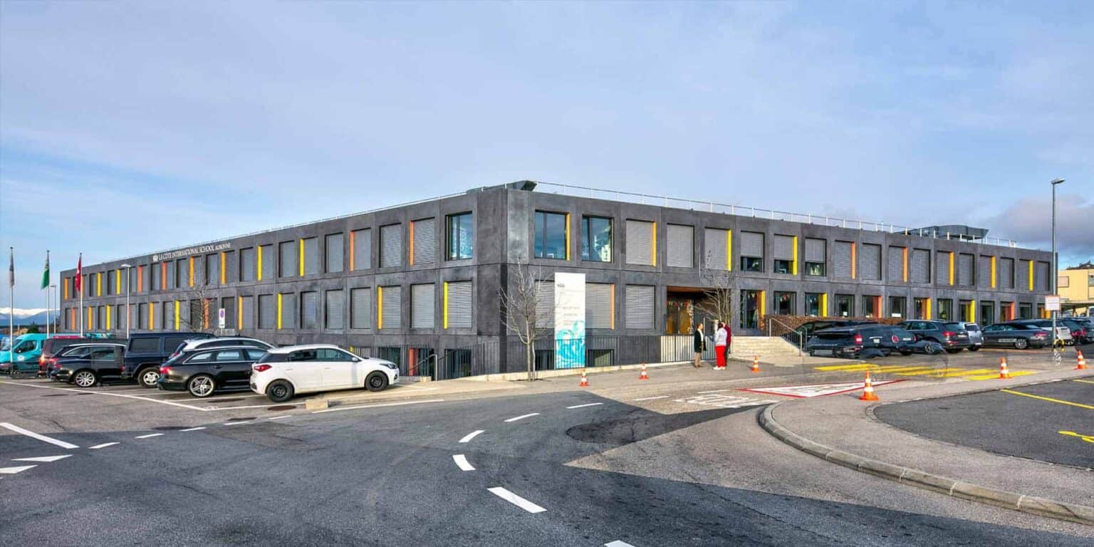 Modern school building with colorful window accents, parking lot, and students entering campus, educational institution for global students.