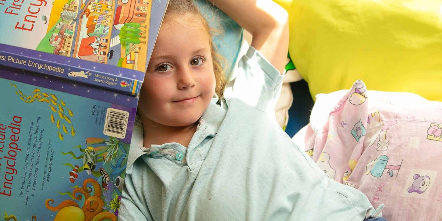 Bright-eyed young girl with books about childhood education and Encyopedia, lying on colorful bedding at school or home, enjoying learning and reading time.
