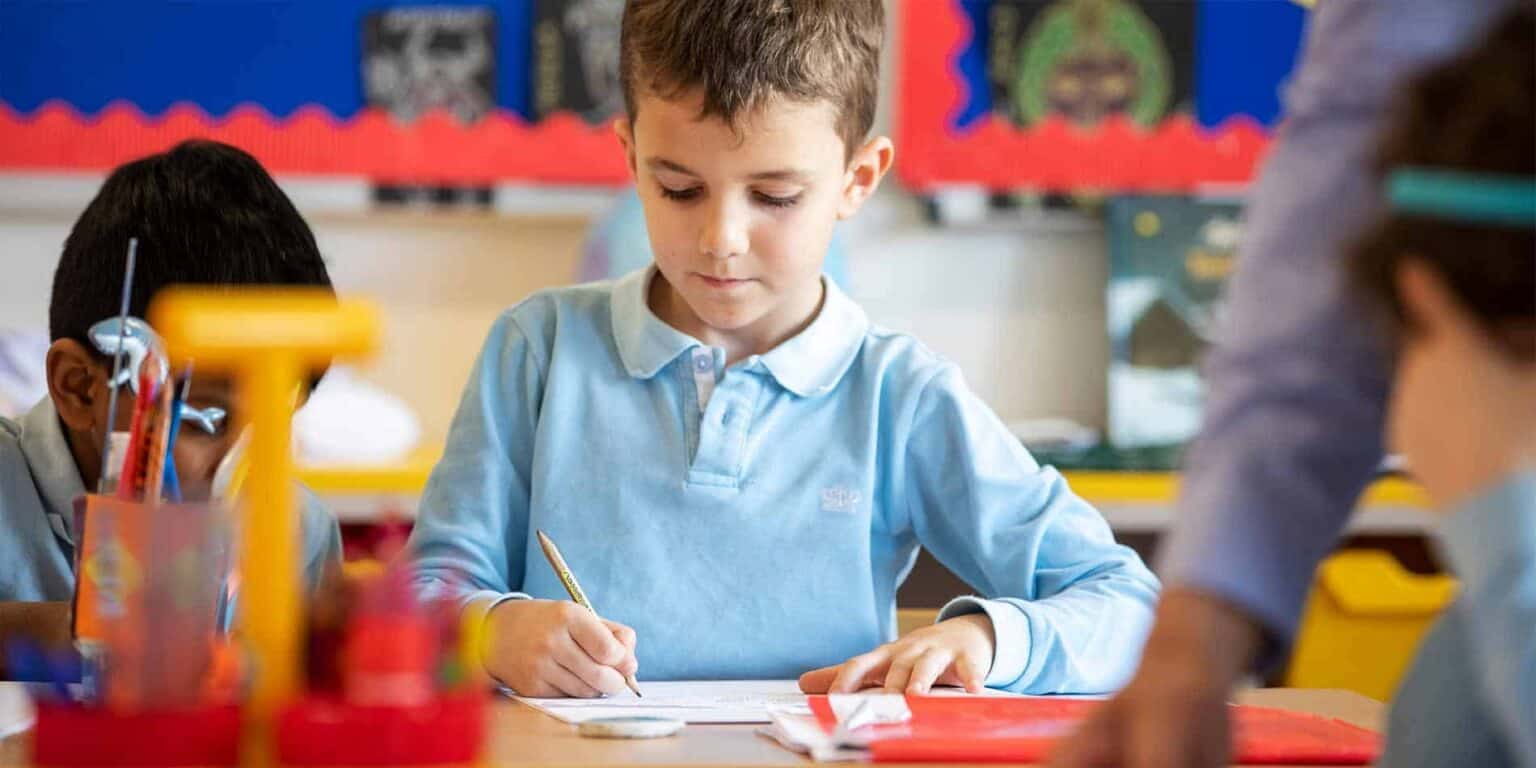 Young boy focused on writing in classroom with educational supplies and colorful decor.