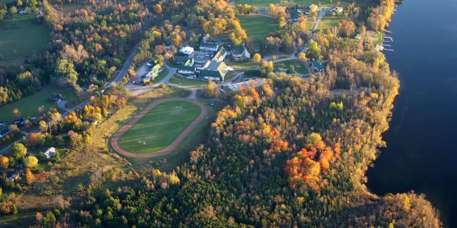 Vista aérea de un pintoresco campus escolar cerca de un lago con follaje otoñal, campos deportivos y un entorno rico en naturaleza.