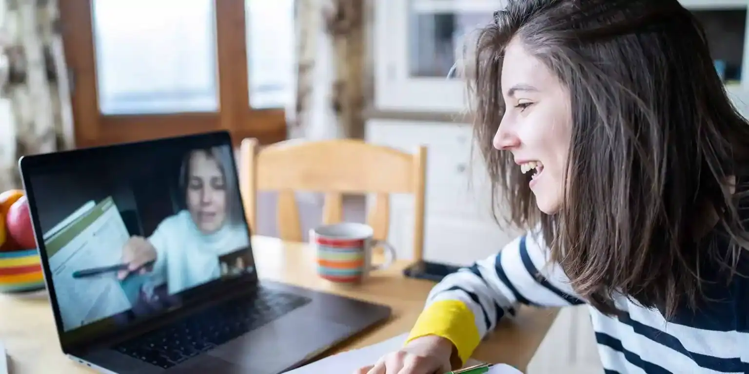 Una joven aprende en línea a través de un ordenador portátil en casa, sonríe e interactúa con el profesor en una videollamada.