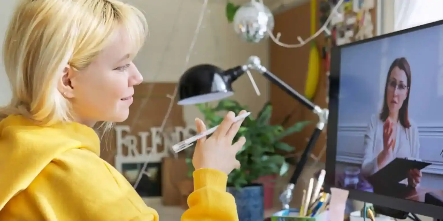 Blonde woman in yellow sweater participating in online learning session with a teacher on video call.