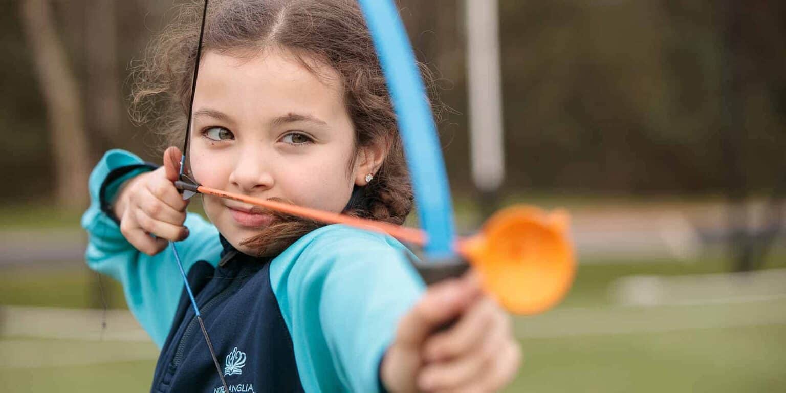 Young girl aiming with bow and arrow during outdoor archery practice at a school sports field.