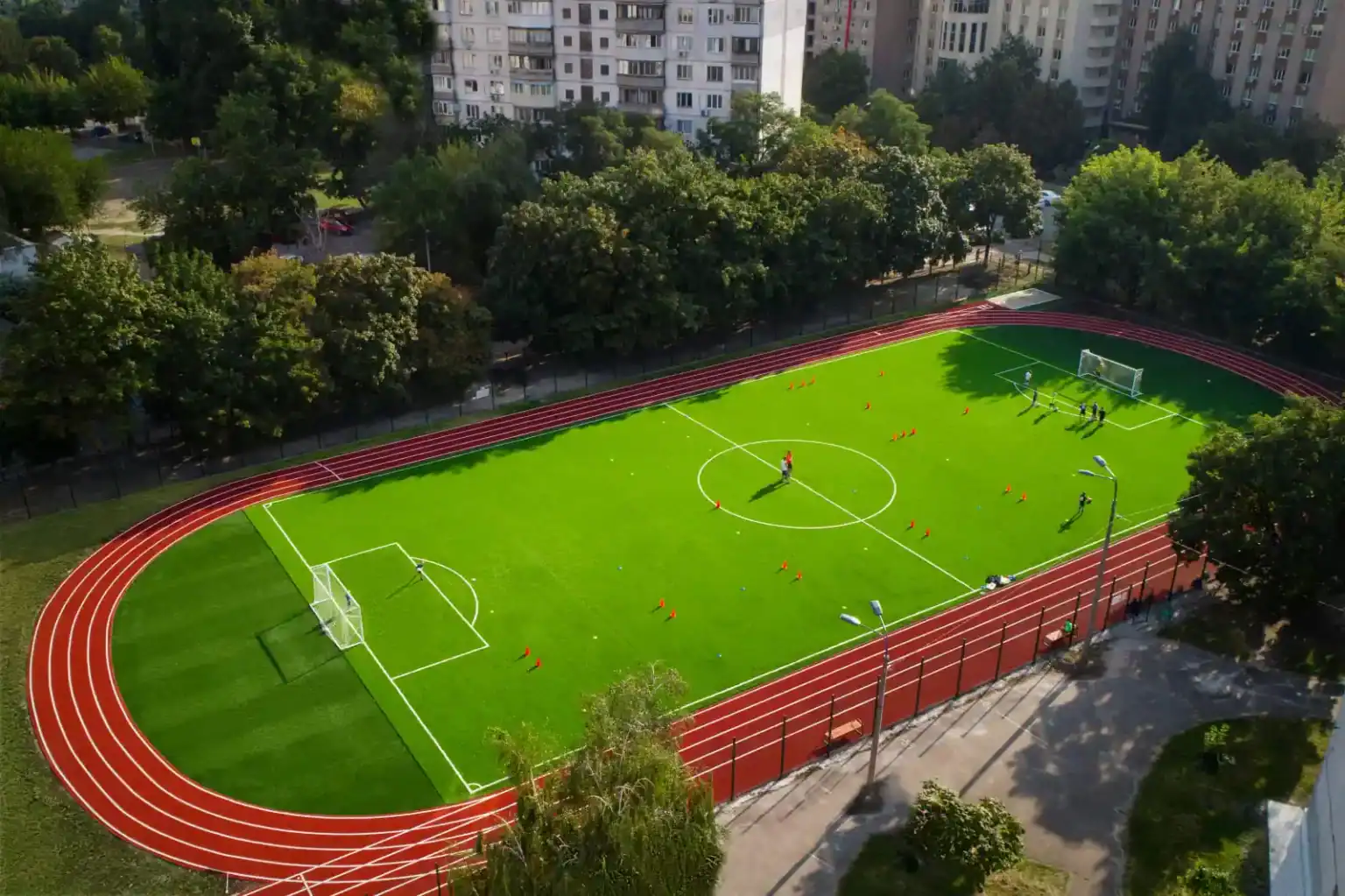 Bright green school sports field with running track, goals, and children practicing soccer in an urban setting.