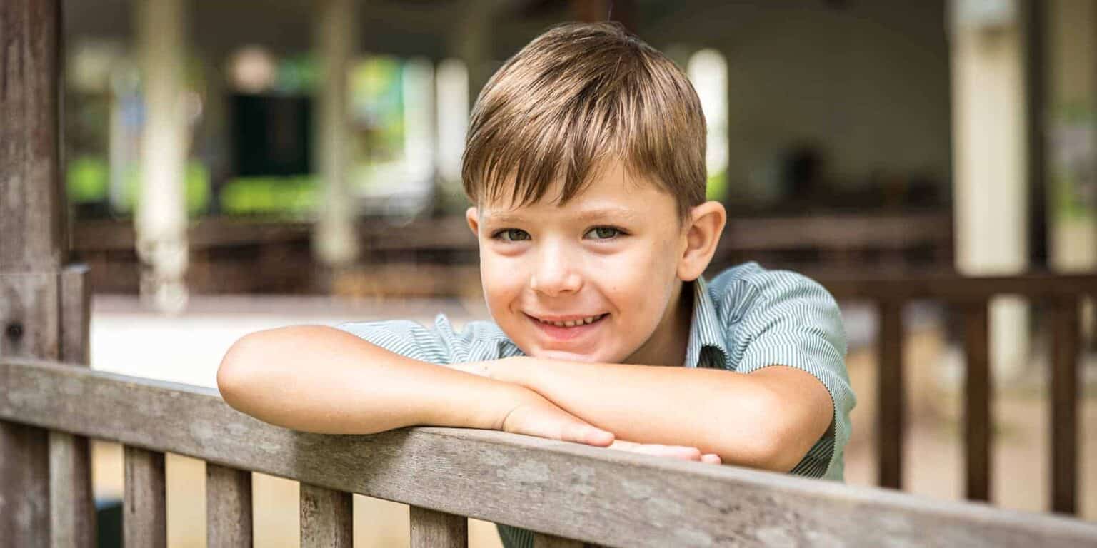 Smiling young boy leaning on a wooden fence at an outdoor school setting, representing quality education and childhood development.