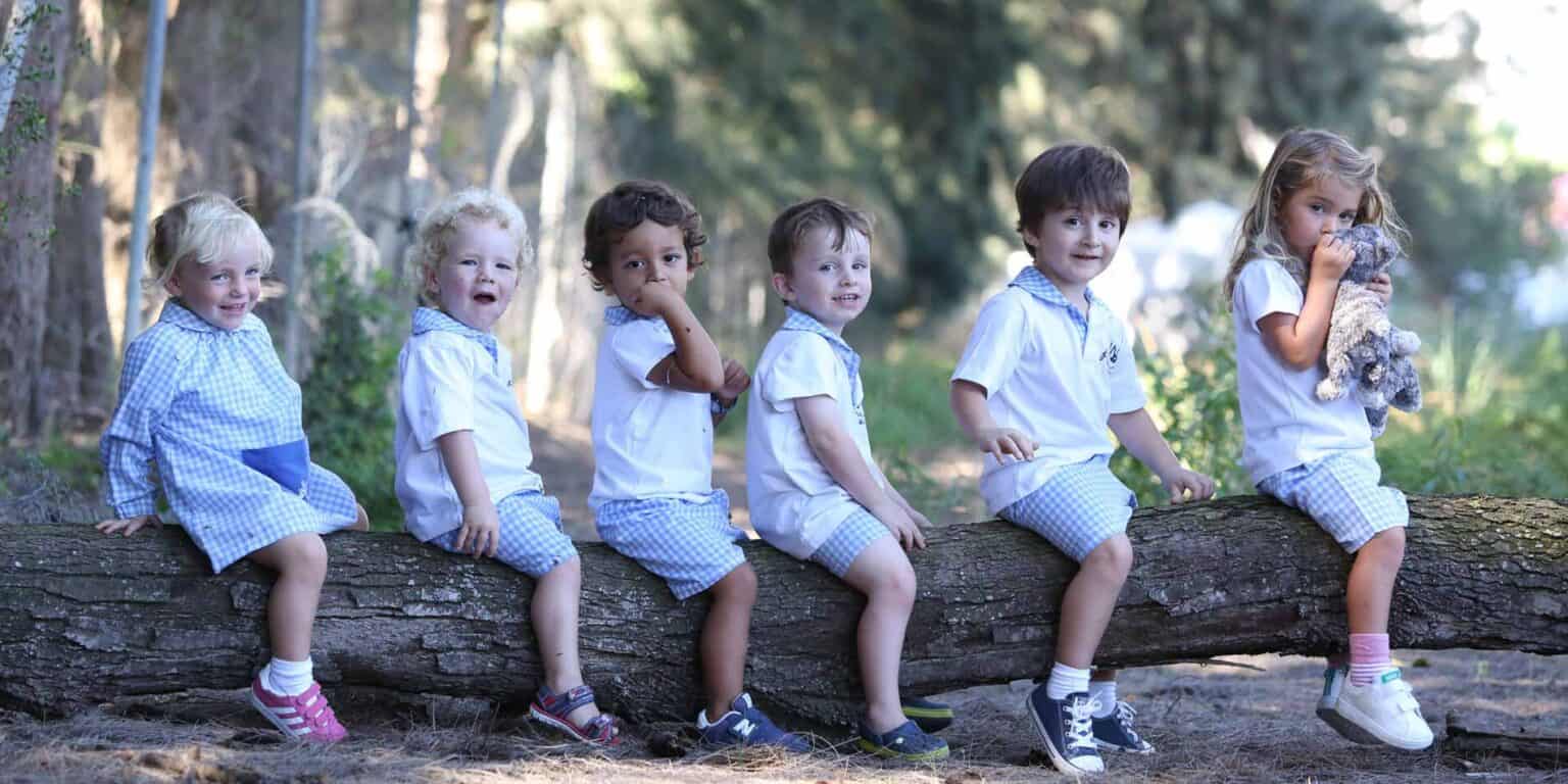 Colorful preschool children sitting on a tree log outdoors in a forest setting.