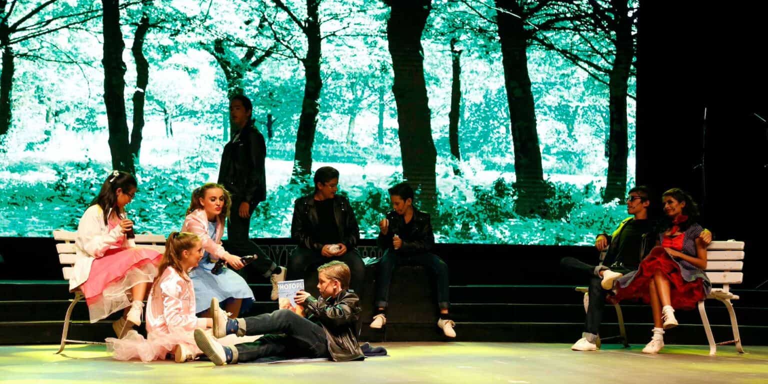 Brightly lit stage with children in vintage costumes, performing in front of a forest backdrop, showcasing a school theater production.