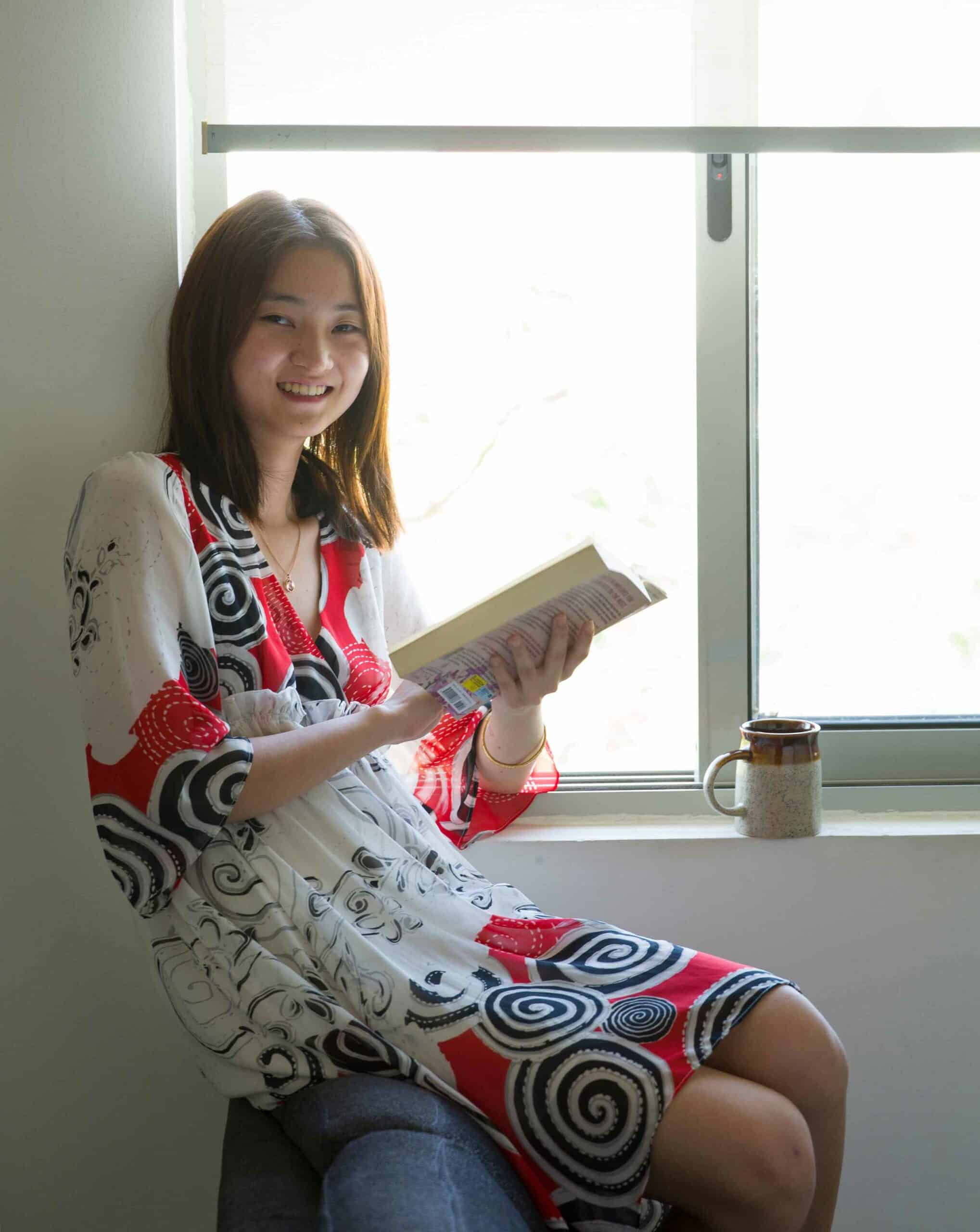 Smiling young woman reading a book by the window at a cozy indoor space, representing academic excellence at World Schools.