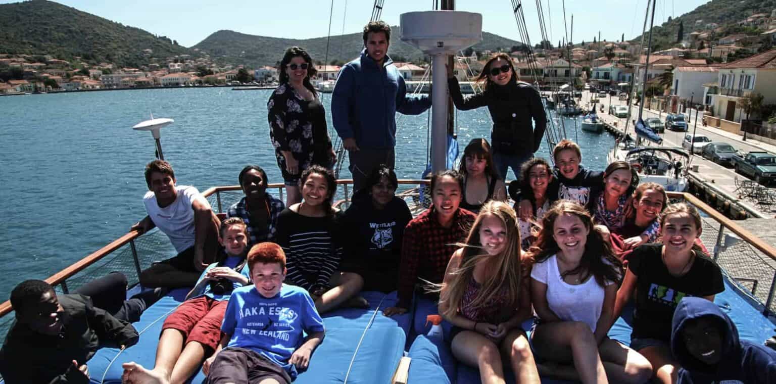 Enjoying a school trip on a boat with diverse students in a scenic harbor setting.