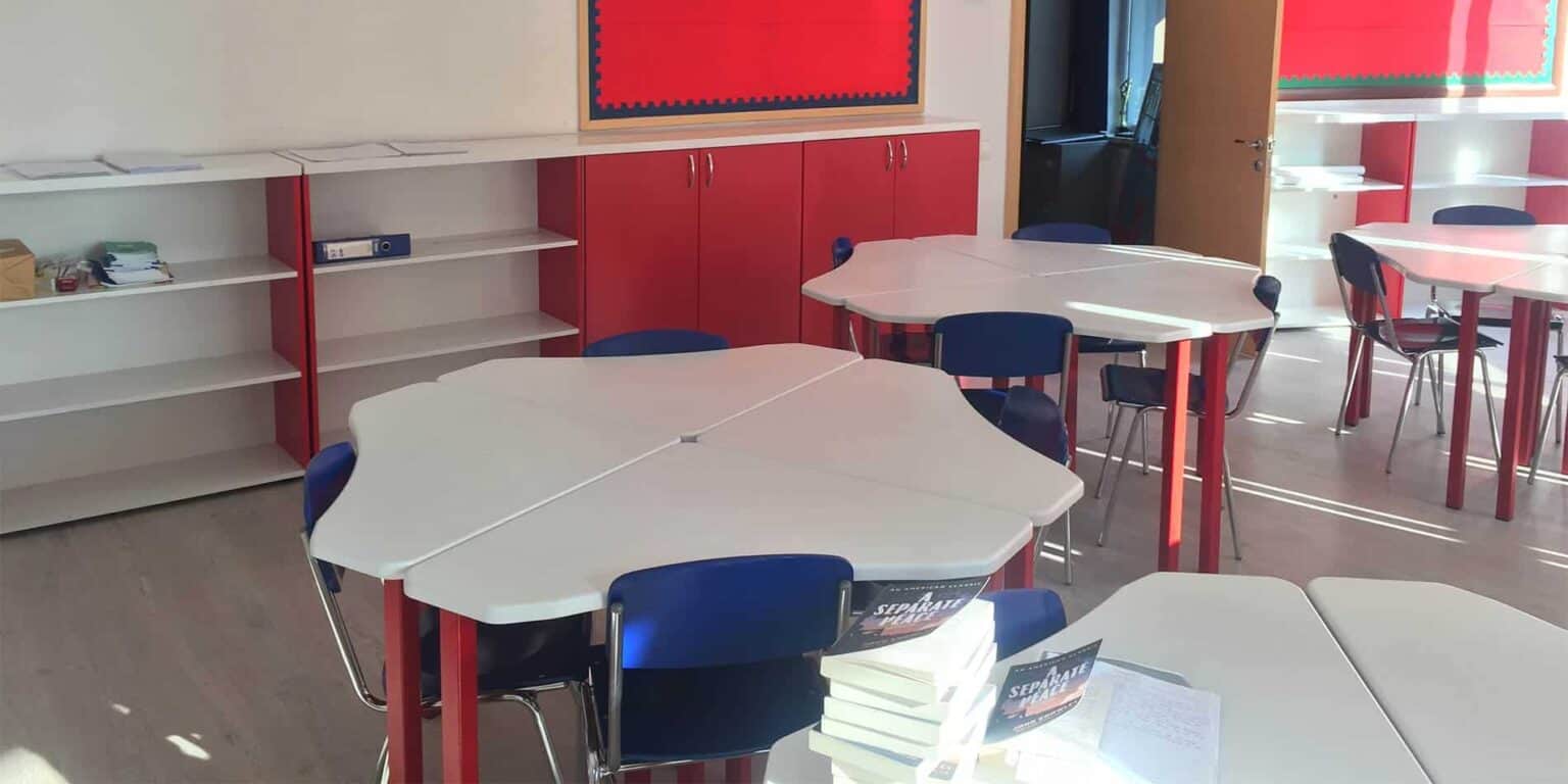Bright classroom with white and red desks and chairs, educational materials, and colorful display boards.