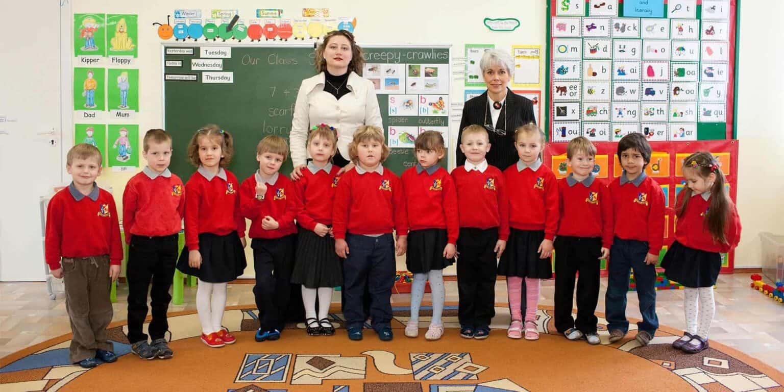 Bright young students in colorful school uniforms pose in classroom with teachers, educational posters, and learning materials.