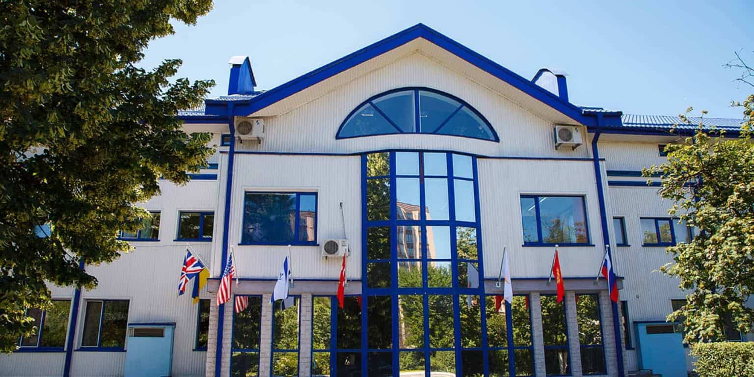 Modern international school building with flags, large glass windows, blue and white exterior for global student community.