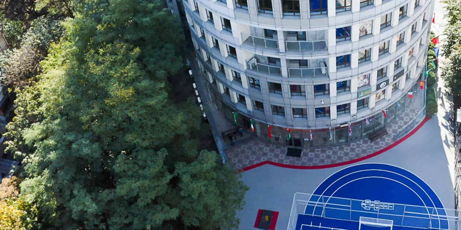 Modern high-rise school building with outdoor sports court and lush green trees.