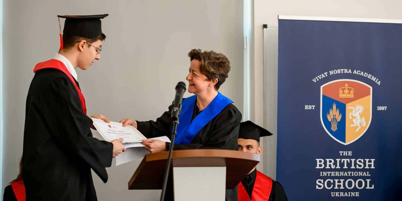 Graduate receiving diploma at school ceremony with teacher congratulating, UK-based international school graduation event.