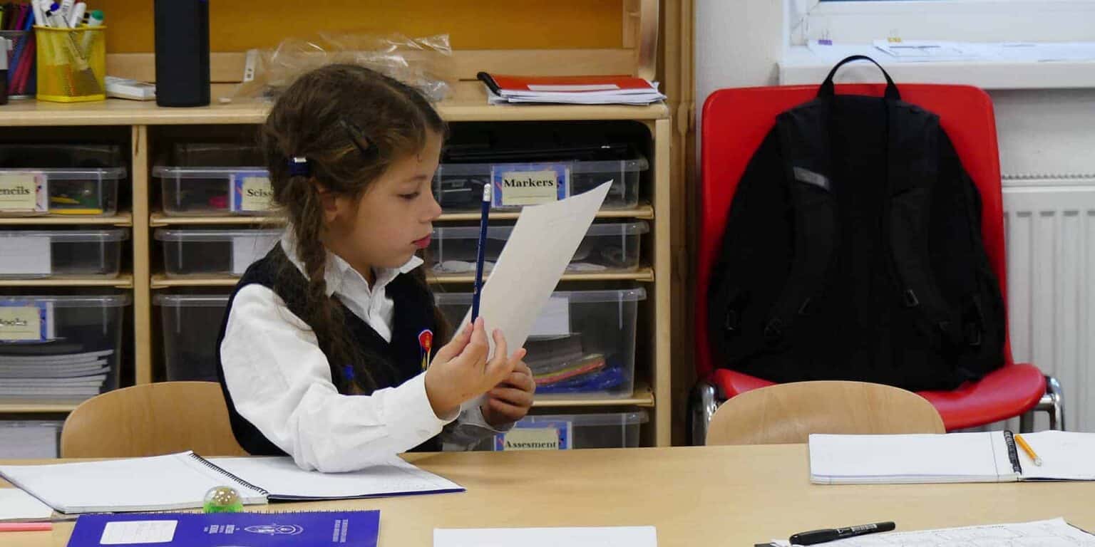 Focused elementary school girl reading a worksheet at school desk with backpacks and classroom supplies.