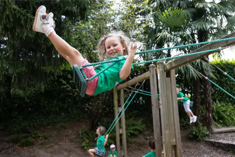 This Month at TASIS: June 2024 31 Children playing on outdoor rope swings at a lush school playground, promoting outdoor activities and physical education.
