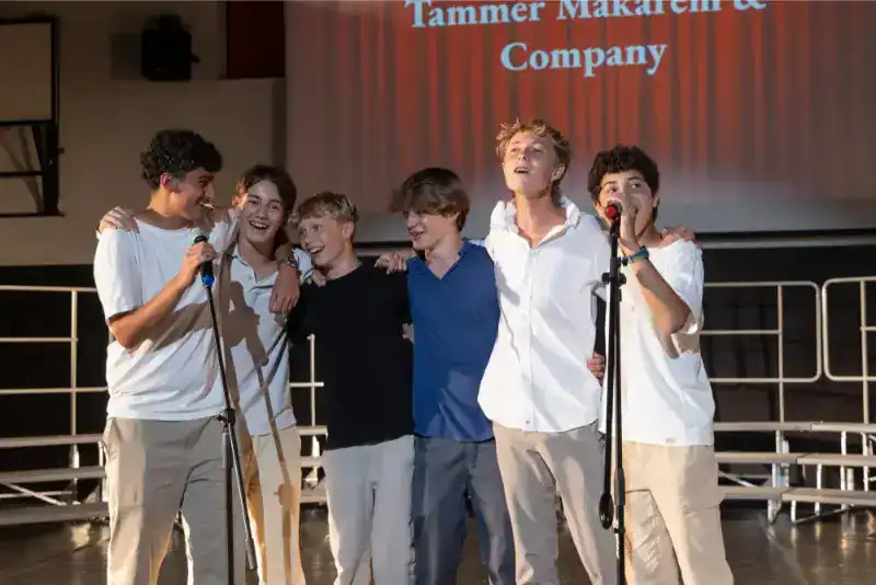 This Month at TASIS: June 2024 39 Happy students singing on stage at a school event with a red curtain backdrop in a modern auditorium.