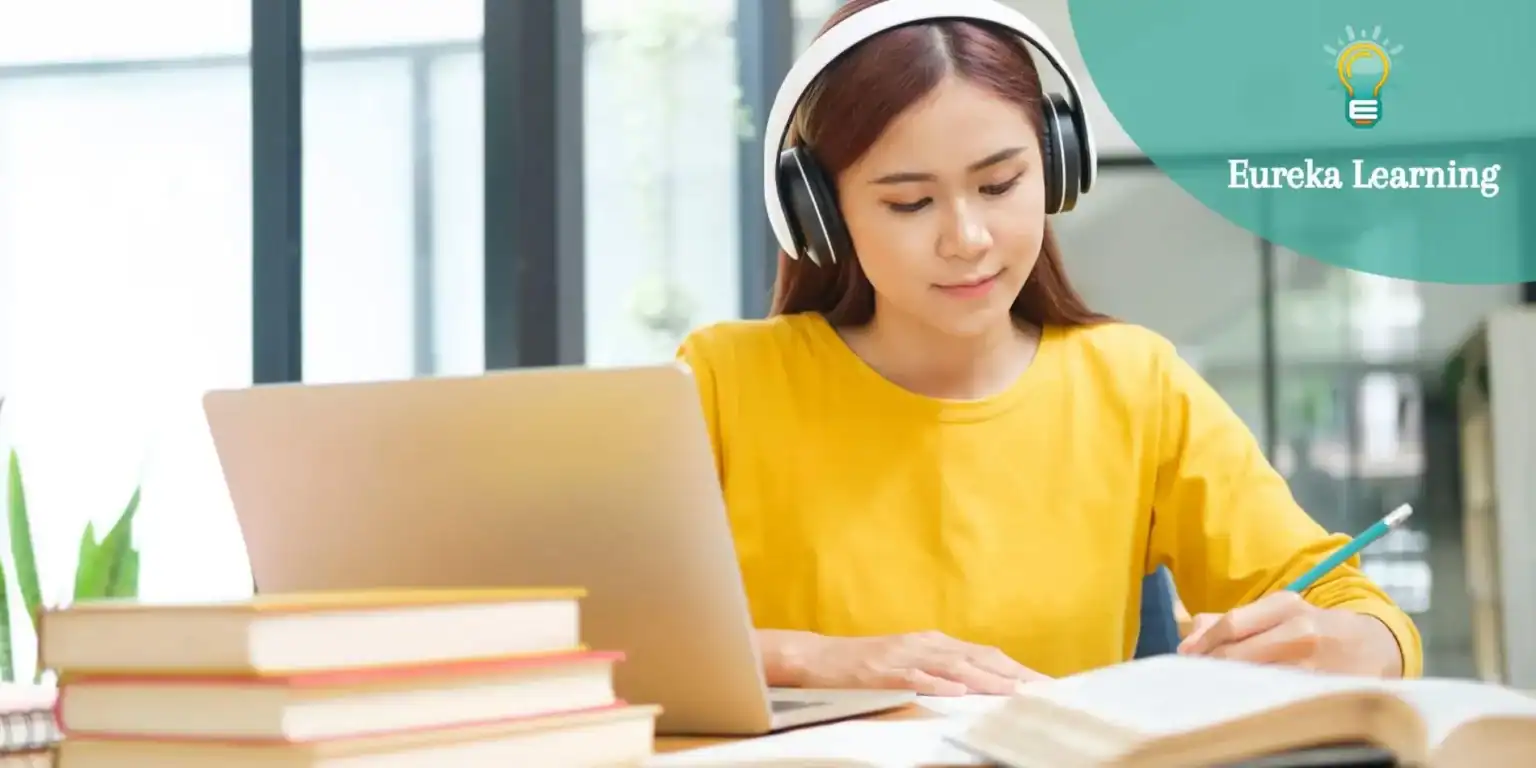 Focused Asian female student studying with headphones, laptop, and books at a modern school or library.