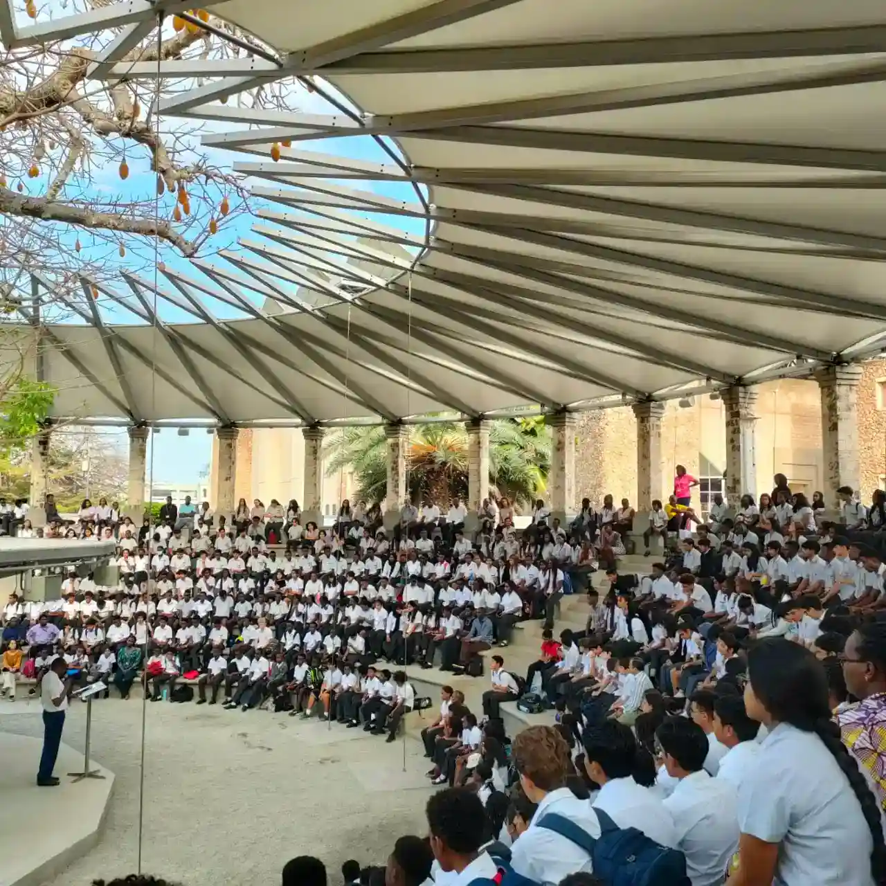 طلاب يحضرون عرضاً كبيراً في الهواء الطلق تحت مدرج حديث مصمم على شكل مظلة في مدرسة عالمية مرموقة.