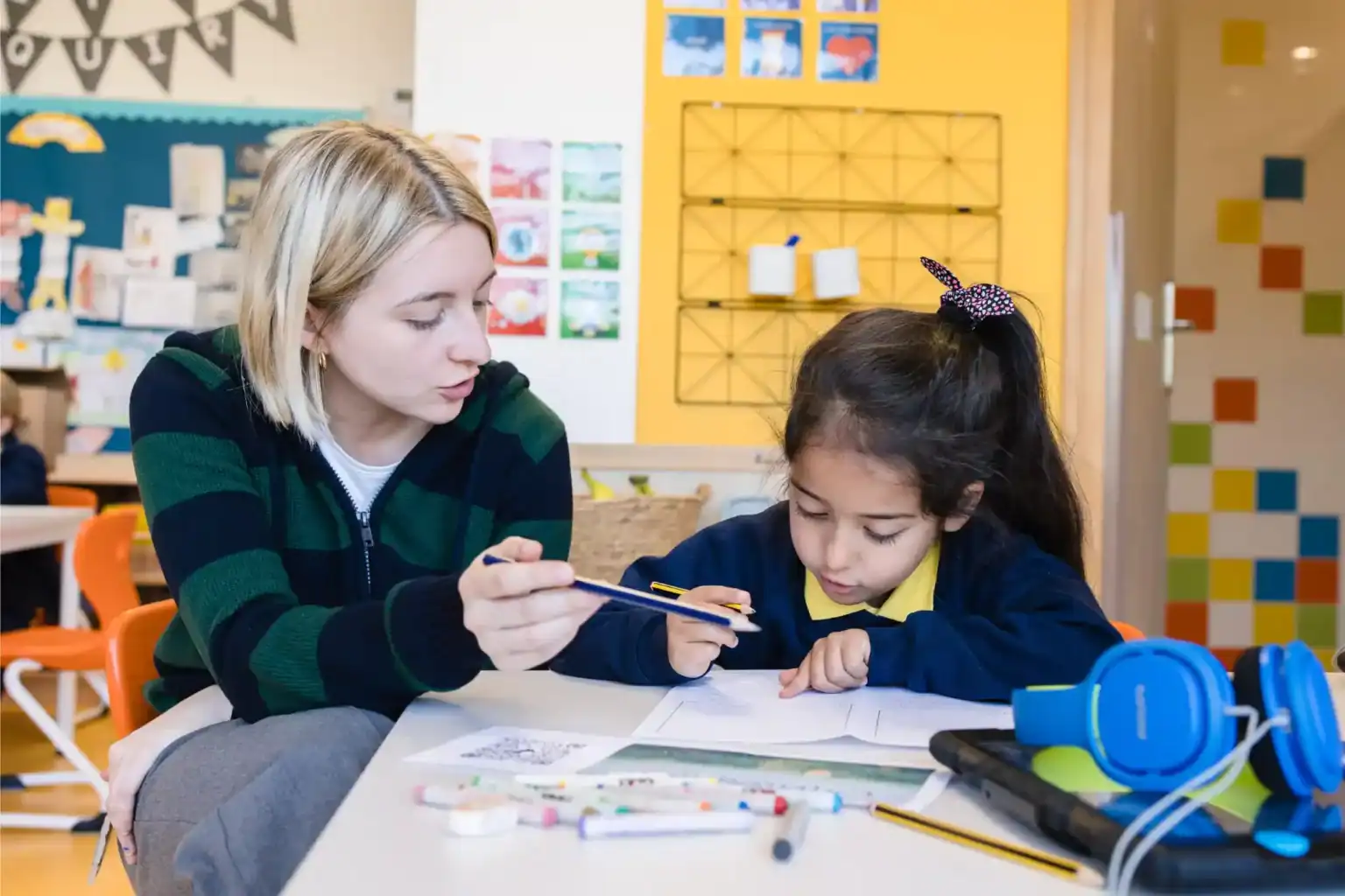 在丰富多彩的教室环境中,专注的老师在辅导小女孩做功课。.