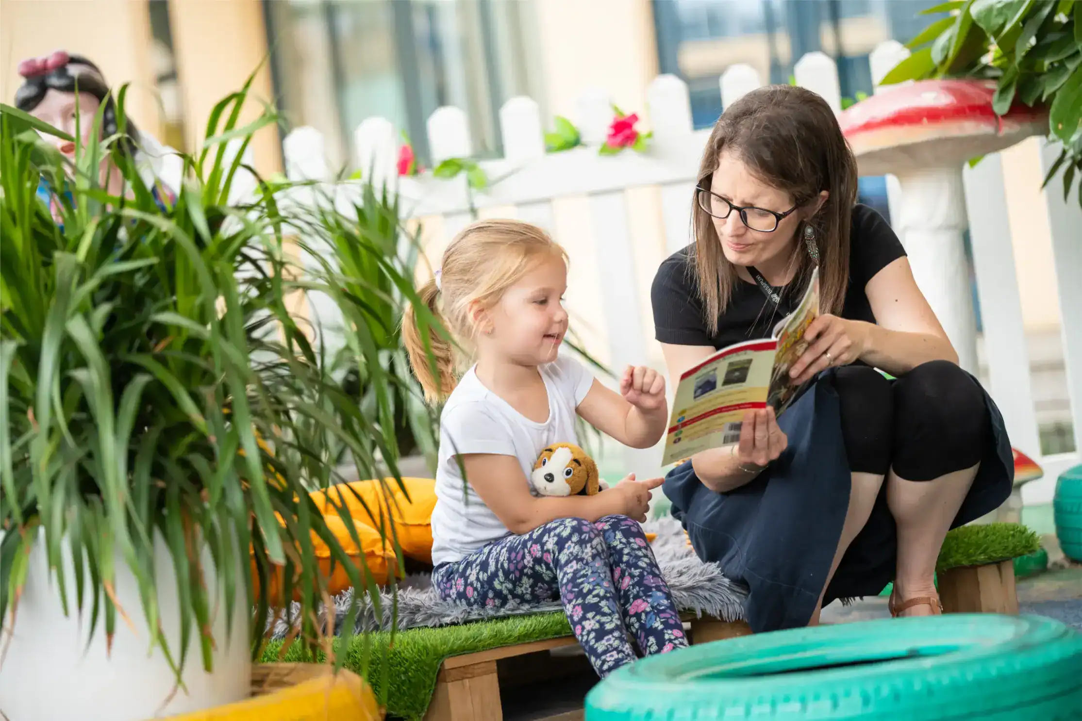 Brightly lit indoor space with a woman and young girl reading together, surrounded by lush green plants and colorful decor.