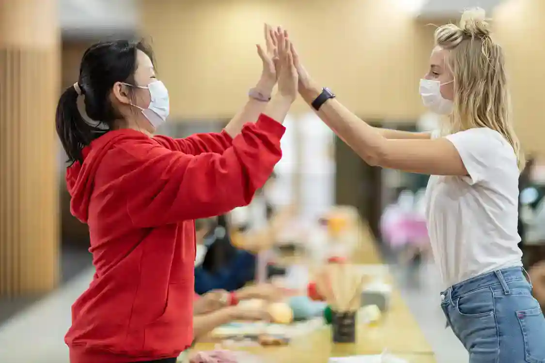 Mask-wearing students giving high-five in school cafeteria for friendly interaction.