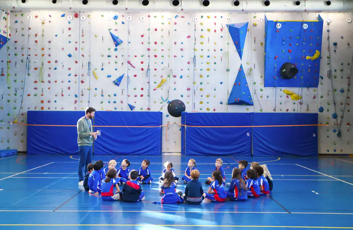 Luminoso rocódromo cubierto en los Colegios del Mundo con jóvenes estudiantes que escuchan las indicaciones del instructor.