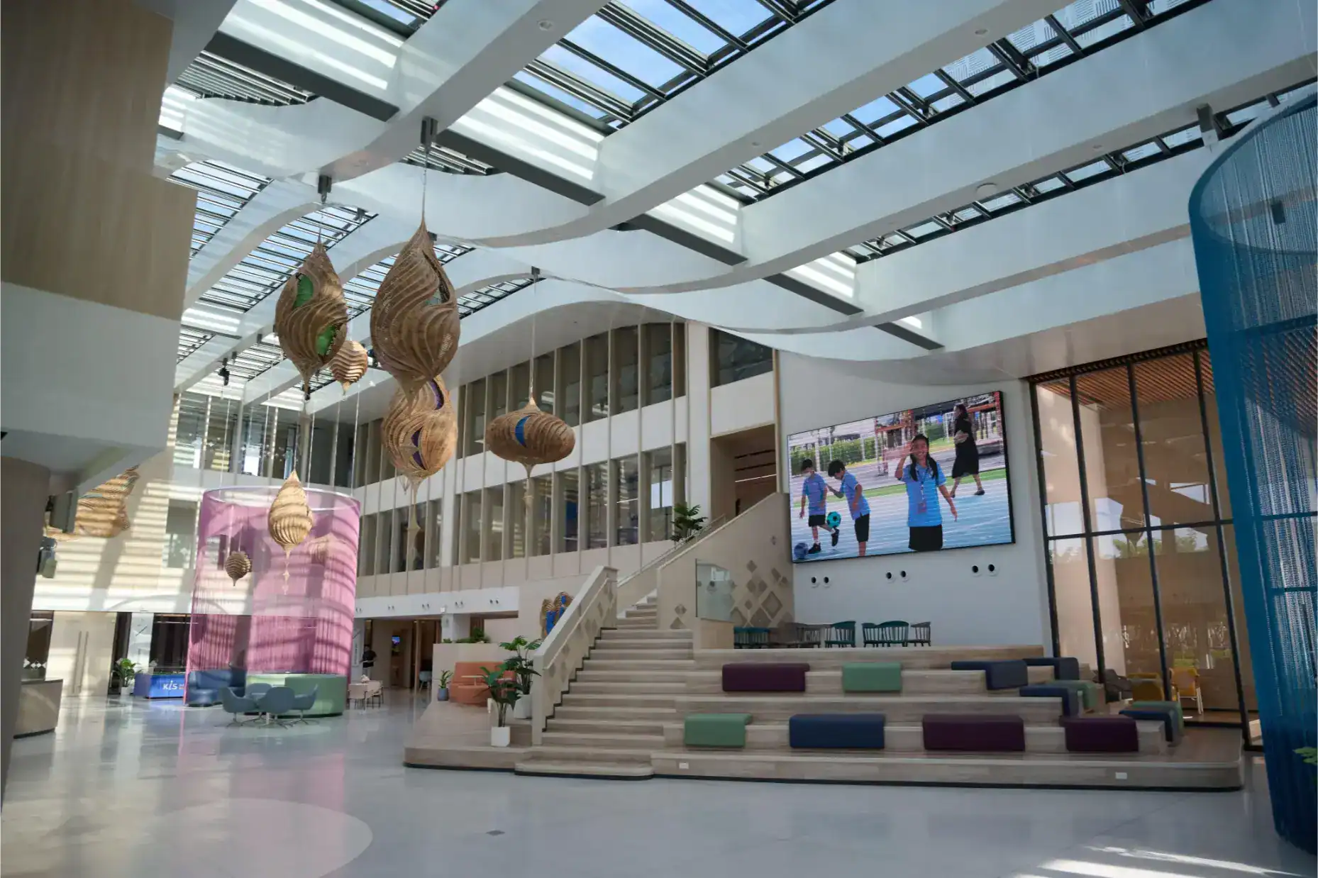 Bright modern school lobby with hanging art installations and large digital screen, promoting innovative education environments.