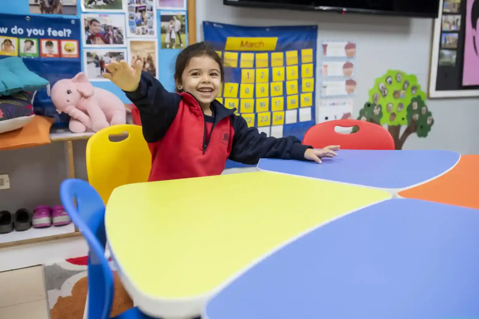 楽しそうに手を振る園児の女の子、カラフルなテーブル、教育的な装飾が施された明るい教室の風景。.