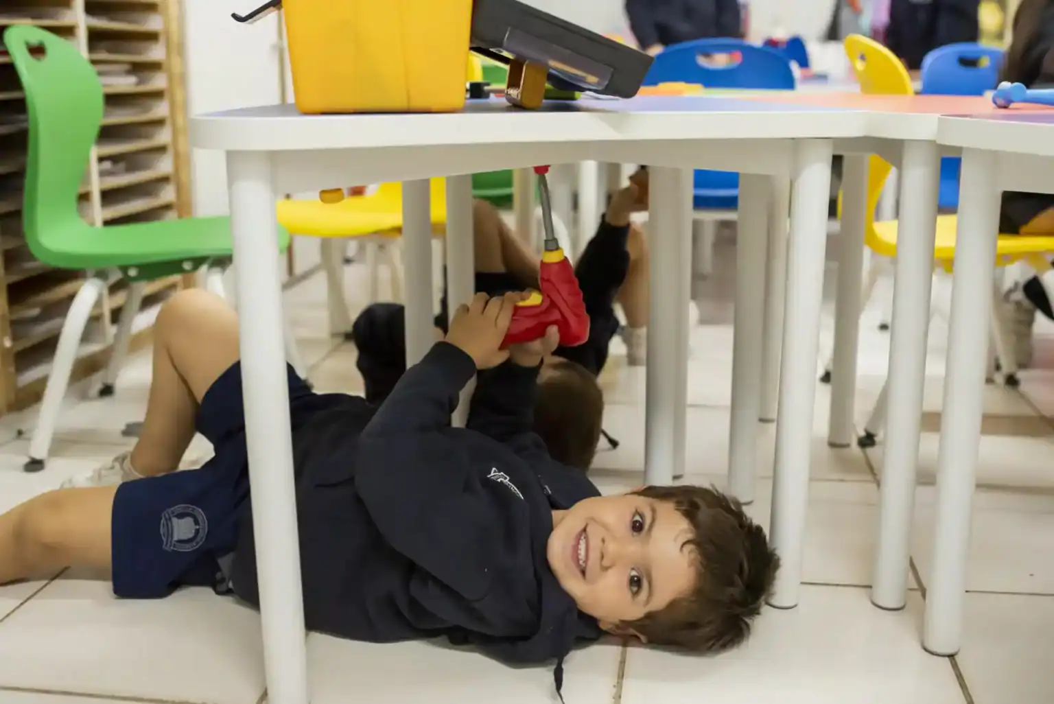 学校の机の下の床に横たわり、おもちゃのドライバーで遊ぶ子供。.