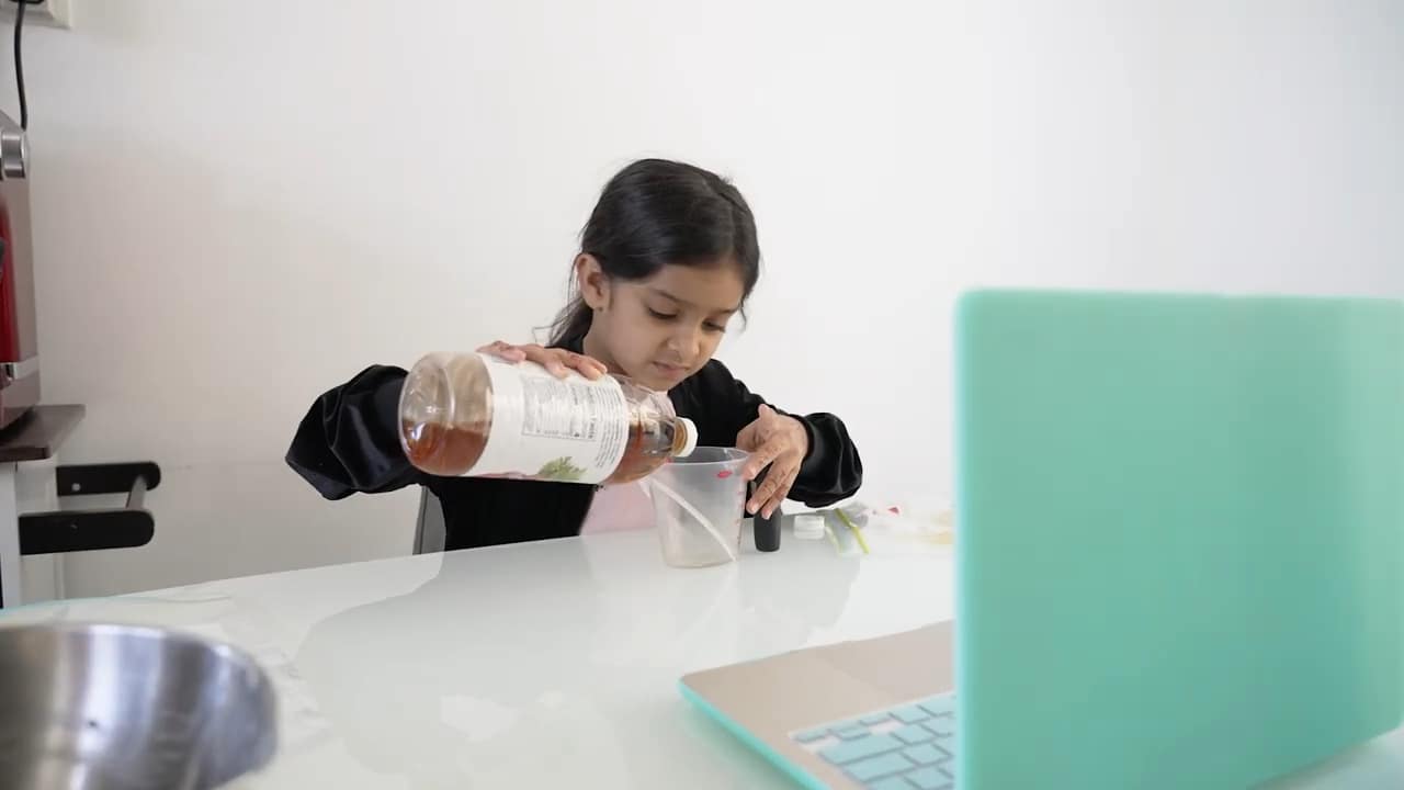 Young girl conducting a science experiment at home, learning chemistry with a measuring cup and laptop.