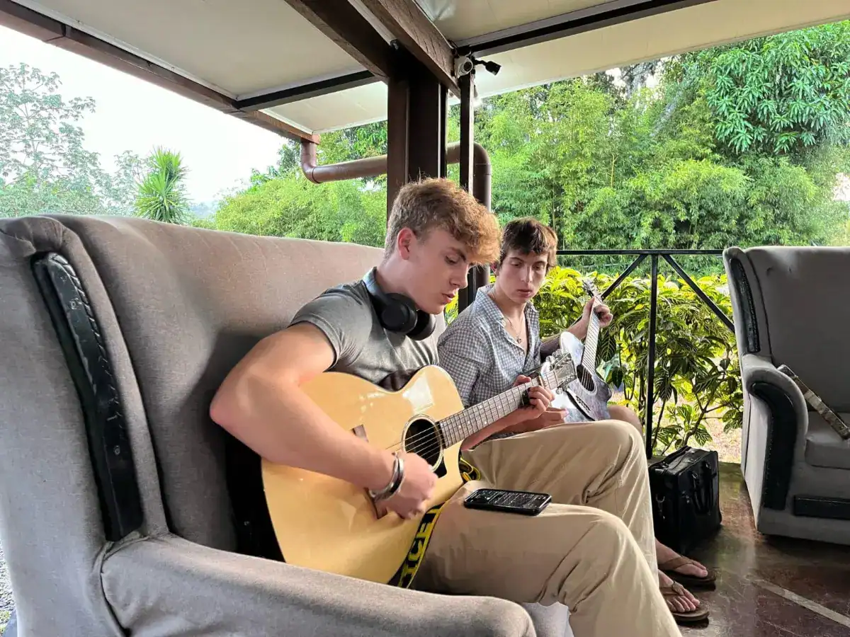 Relaxed teenage boys playing acoustic guitars on outdoor patio, green lush landscape background, leisure, music, youth culture.