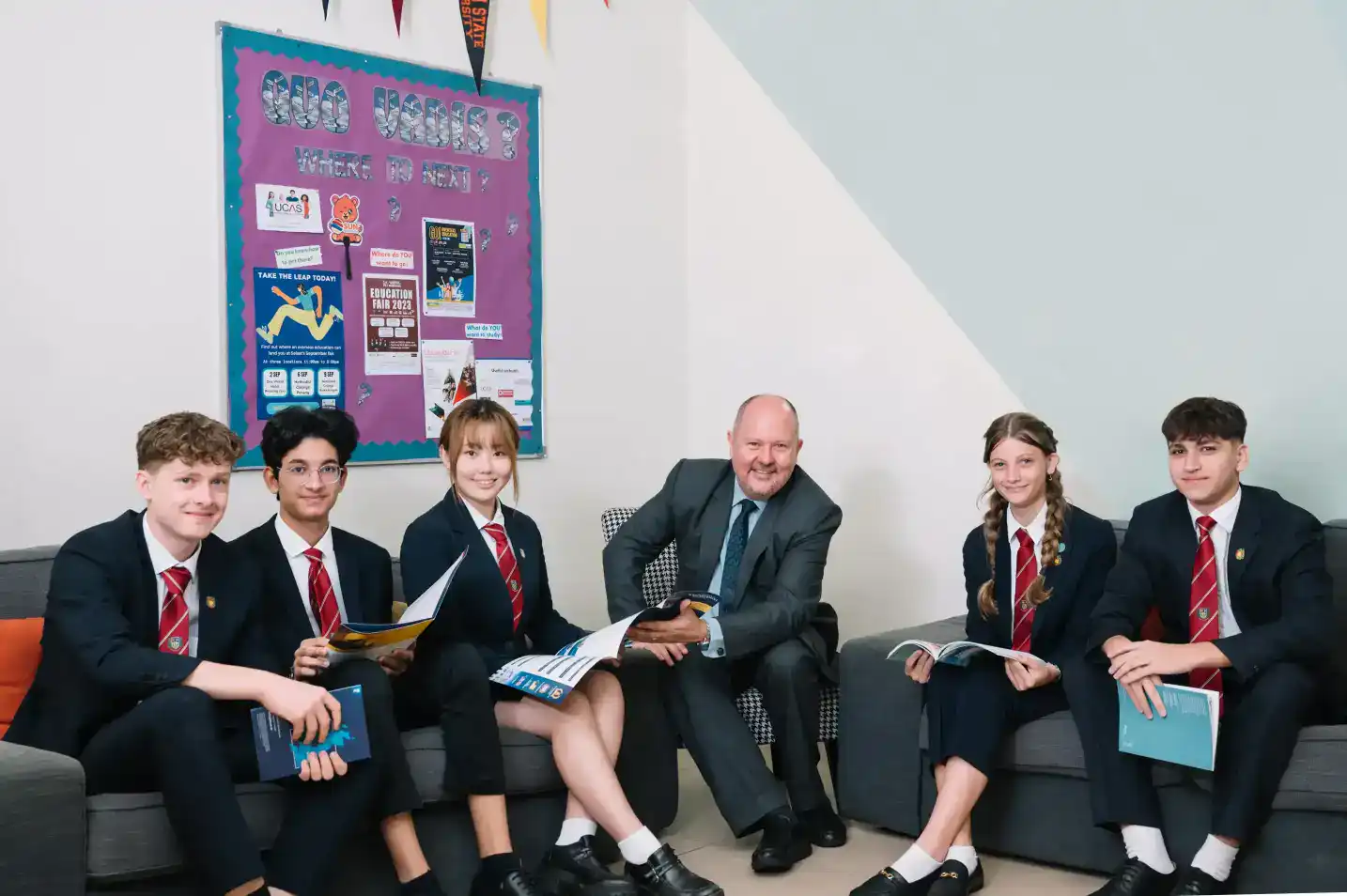 Students in school uniforms engaged in discussion with a teacher in a classroom setting.