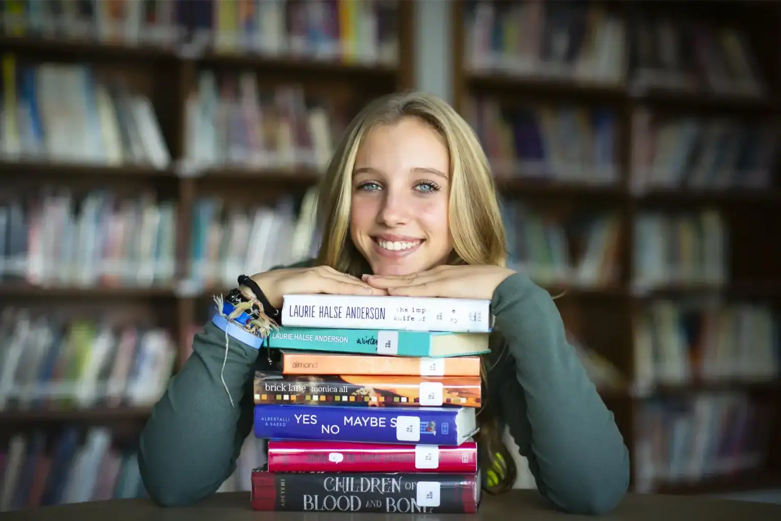 Young smiling girl holding a stack of books in a school library, promoting education and literacy.