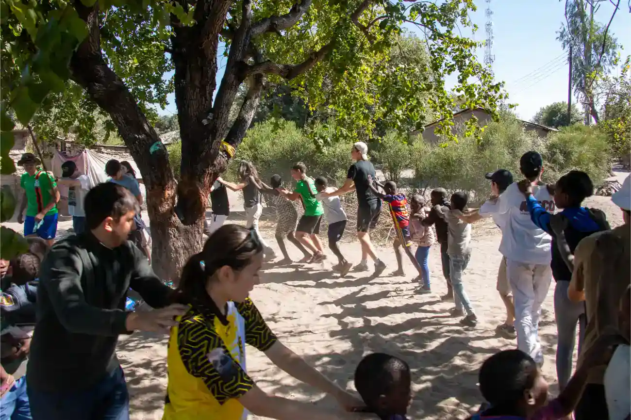 This Year at TASIS: 2023 25 Children playing tug of war outdoors at a school activity under a large tree in a rural setting, multicultural students having fun, community event.