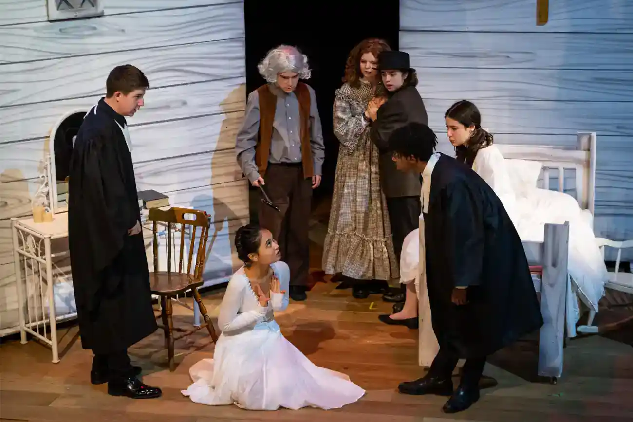 This Year at TASIS: 2023 45 Girl on stage in white dress sitting on floor with people around her.