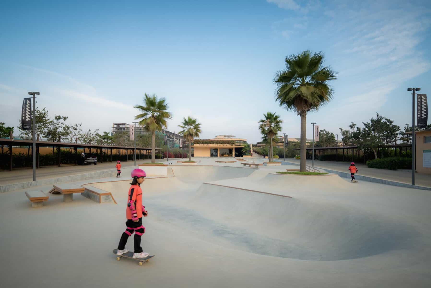 Skateboarders at an outdoor skatepark with palm trees and modern buildings in the background.