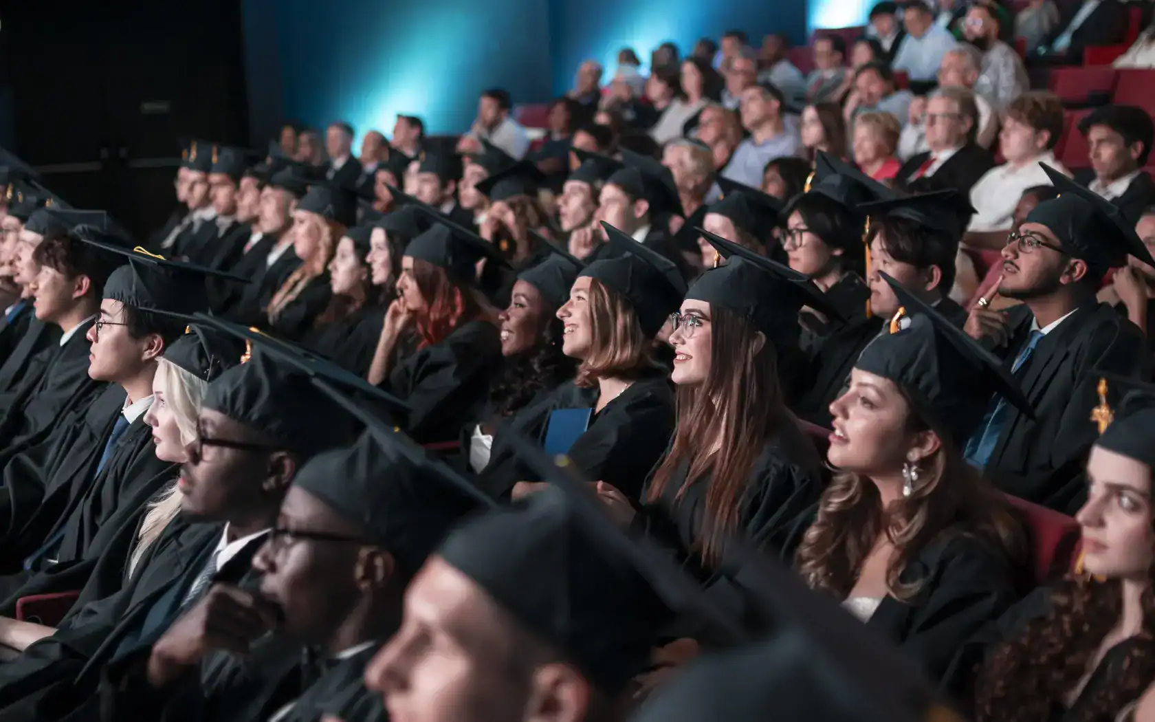 Graduates in caps and gowns attending a formal school graduation ceremony at a large auditorium.