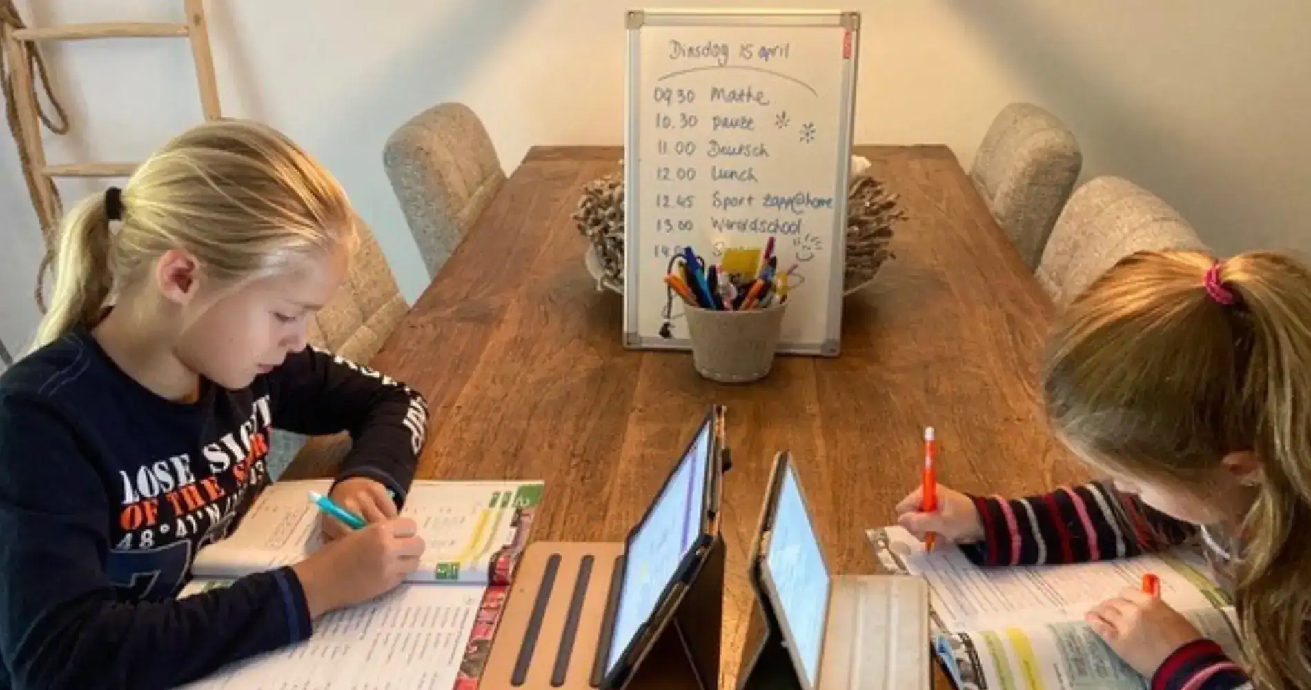 Busy children studying with tablets and books at a wooden table in a home or classroom setting.