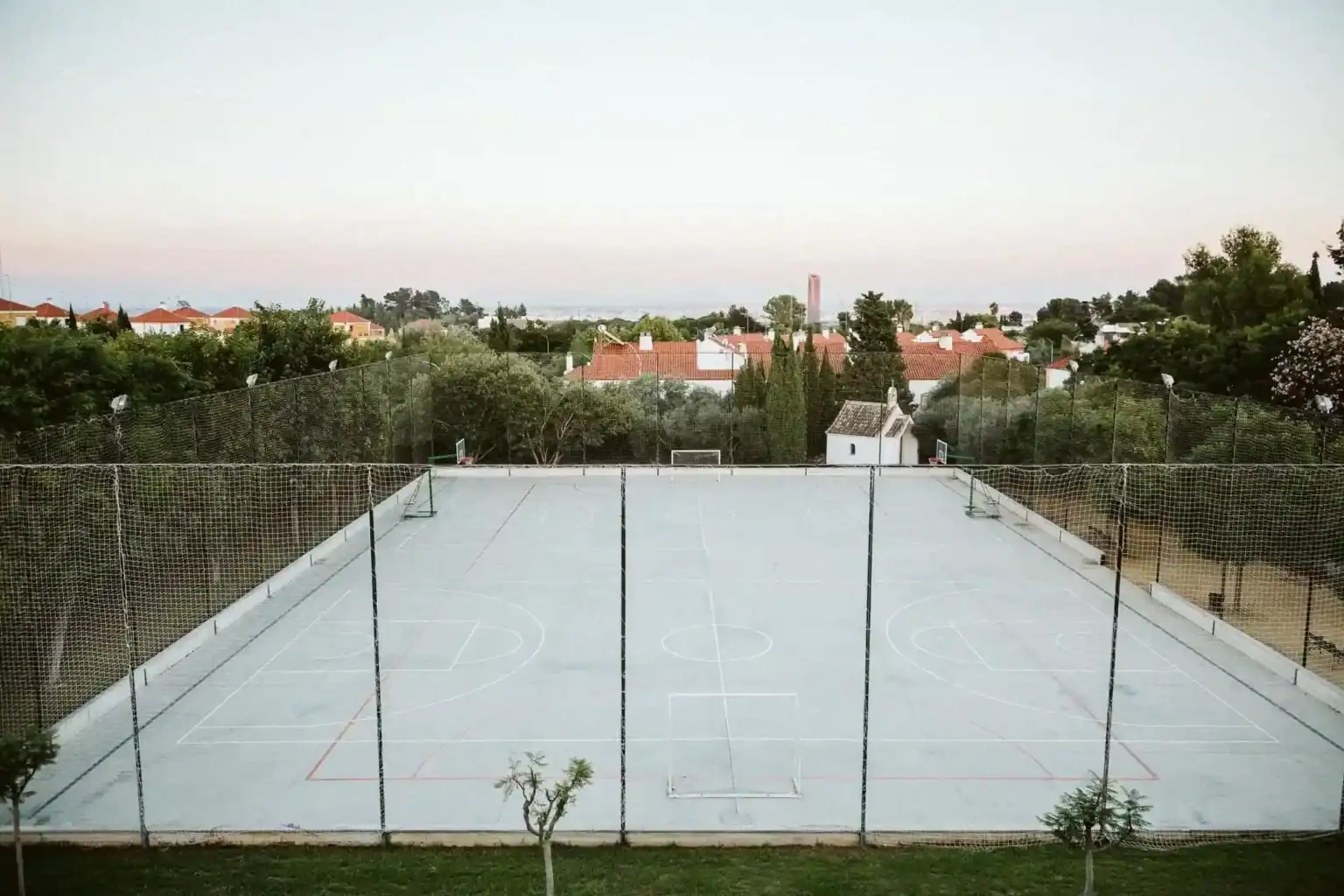 Outdoor school basketball court surrounded by trees and residential houses, ideal for student sports activities.