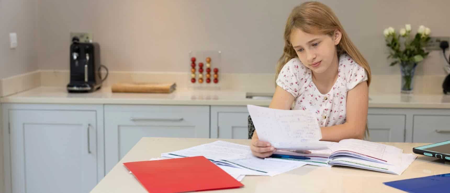 Young girl doing homework at kitchen table with notebooks and papers, studying for school.