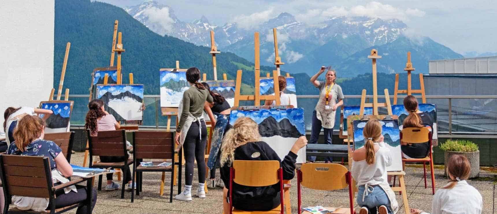Colorful outdoor painting class at a world school in scenic mountain setting with students and teacher.