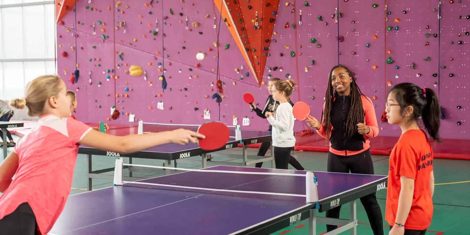 Girls playing table tennis in a school sport hall with climbing wall on background.